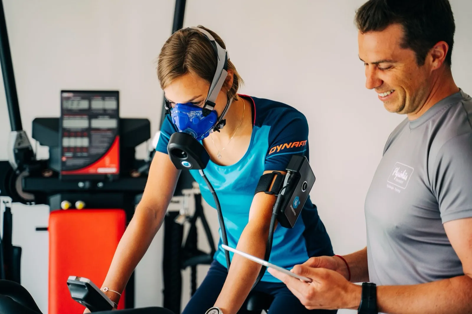 Eine Athletin fährt unter Belastung auf dem Ergometer und trägt dabei eine Atemmaske zur Messung der Atemgase; daneben liest ein Therapeut von physio4me die Werte auf dem Tablet ab.
