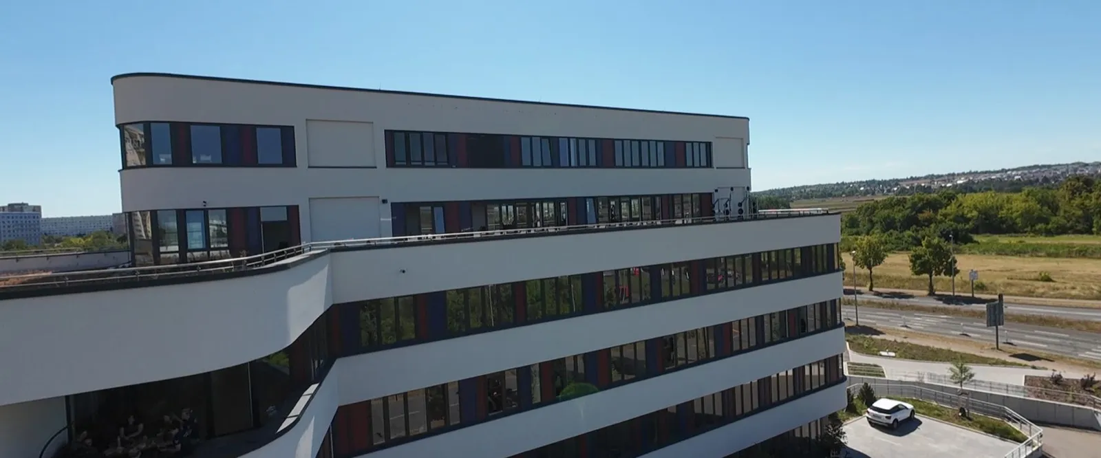 Luftaufnahme des Gebäudes der Polyklinik Nord: mehrstöckiger heller Neubau mit Terrassen und rot-blau akzentuierten Fensterbändern unter blauem Himmel.